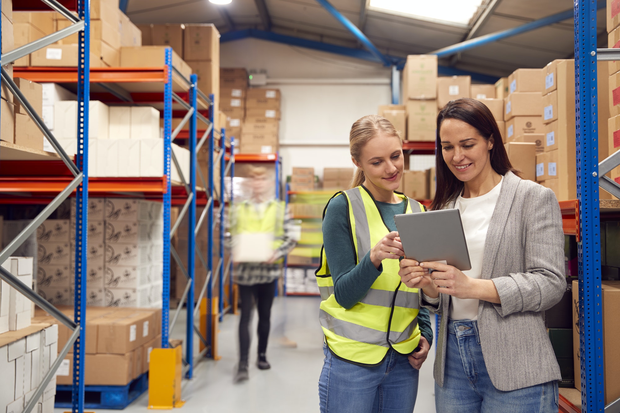 Manager Using Digital Tablet With Female Intern Inside Busy Warehouse Facility